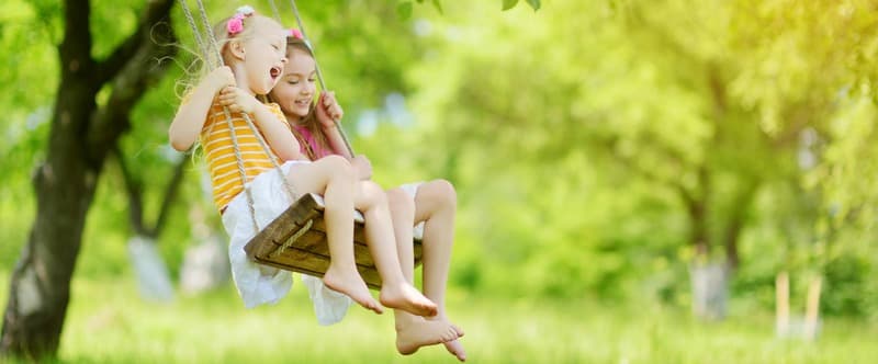 Two cute little sisters having fun on a swing together in beautiful summer garden on warm and sunny day outdoors