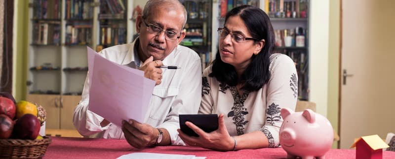 Senior Indian/asian couple accounting, doing home finance and checking bills with laptop, calculator and money while sitting on sofa/couch at home