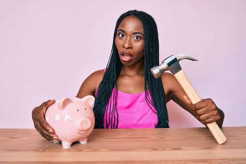 African american woman with braids holding piggy bank and hammer in shock face, looking skeptical and sarcastic, surprised with open mouth